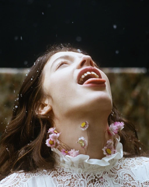 A woman lies on her back with her mouth open, sticking her tongue out. She is surrounded by small flowers and there are dark background.