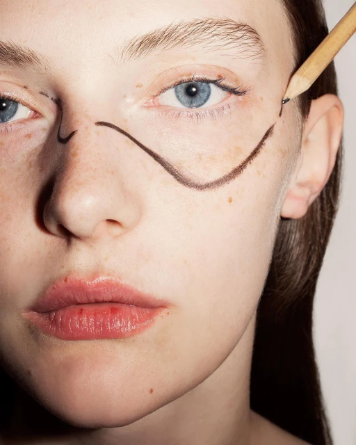 Close-up shot of a fair-skinned woman with freckles and blue eyes, featuring drawn lines on her face and a pencil touching her skin.