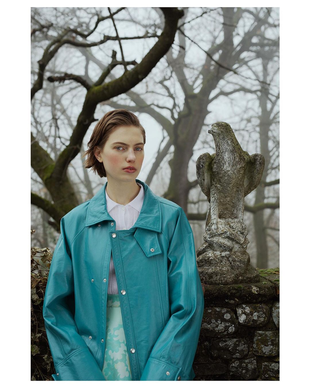 A young woman poses in a teal jacket with a light skirt in an outdoor environment with stone statue and bare trees in soft light.