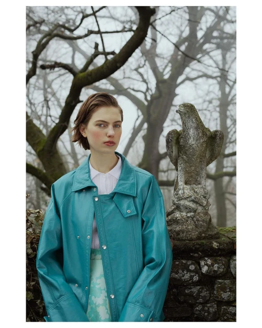 A young woman poses in a teal jacket with a light skirt in an outdoor environment with stone statue and bare trees in soft light.