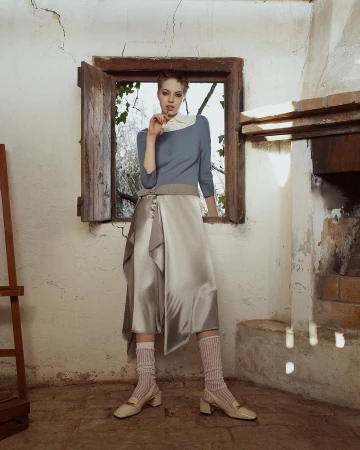 A short-haired woman stands in a rustic room near a wooden window opening, wearing a blue sweater and a satin skirt.