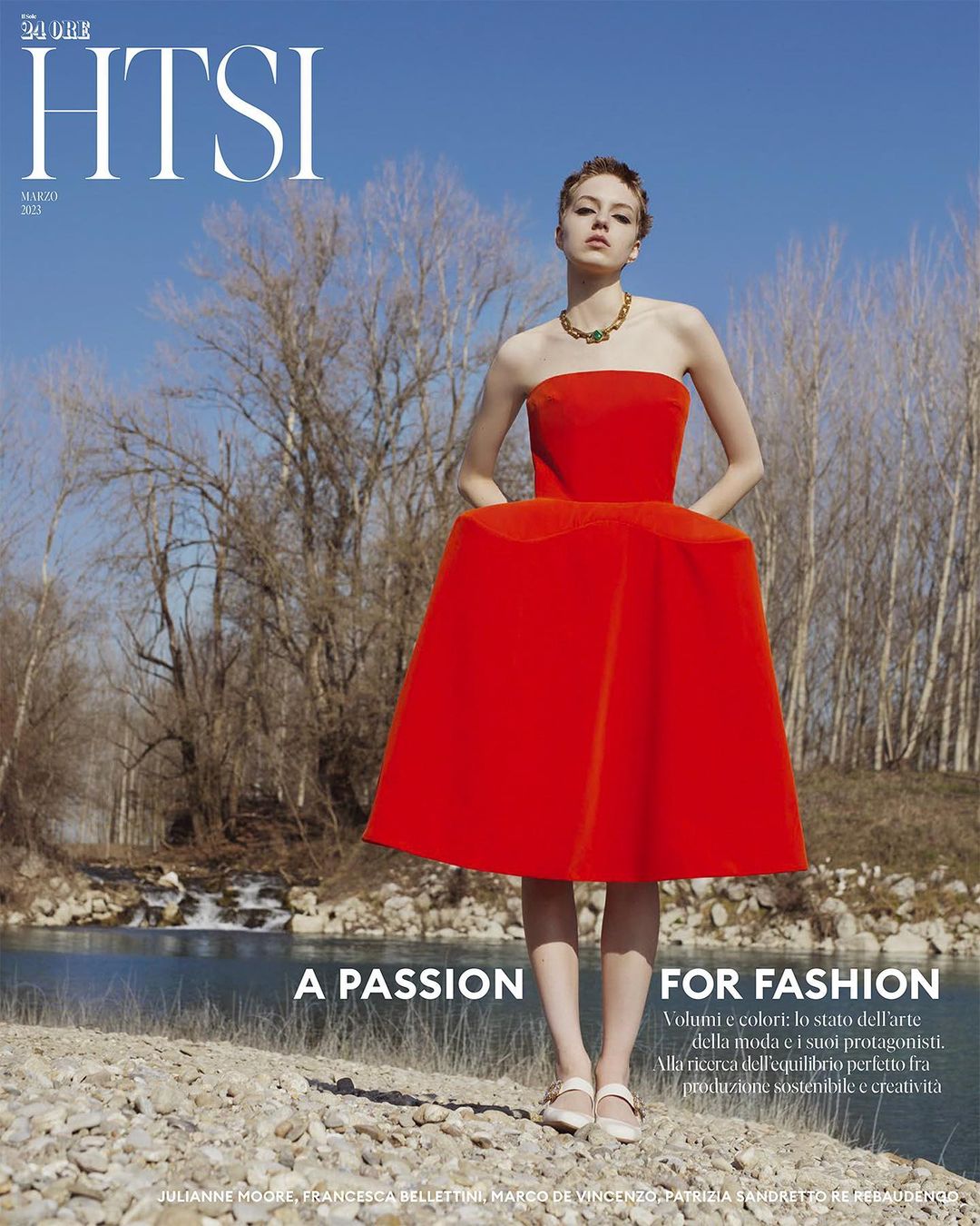 Woman in a vibrant red strapless dress standing on a pebbled riverbank with bare trees against a blue sky.