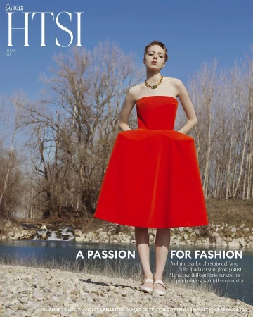 Woman in a vibrant red strapless dress standing on a pebbled riverbank with bare trees against a blue sky.
