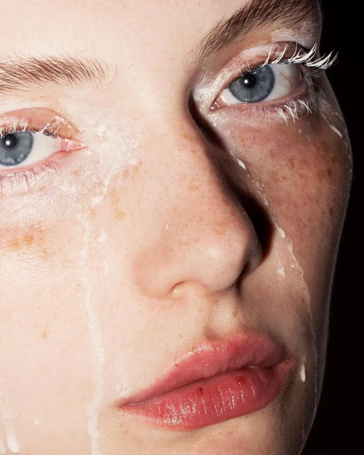 Close-up portrait of a fair-skinned adult female with striking blue eyes, freckles, white eyelashes, and artistic white paint dripping down her face.