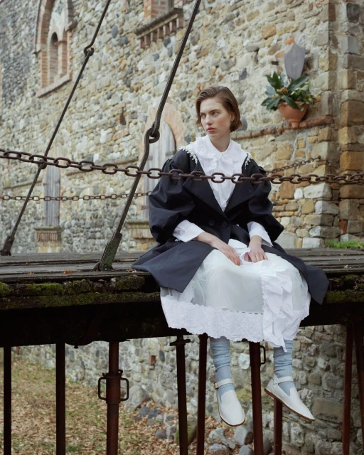 A woman sits on a wooden bridge in a black coat and white dress, with a stone building in the background. Fashion and style.