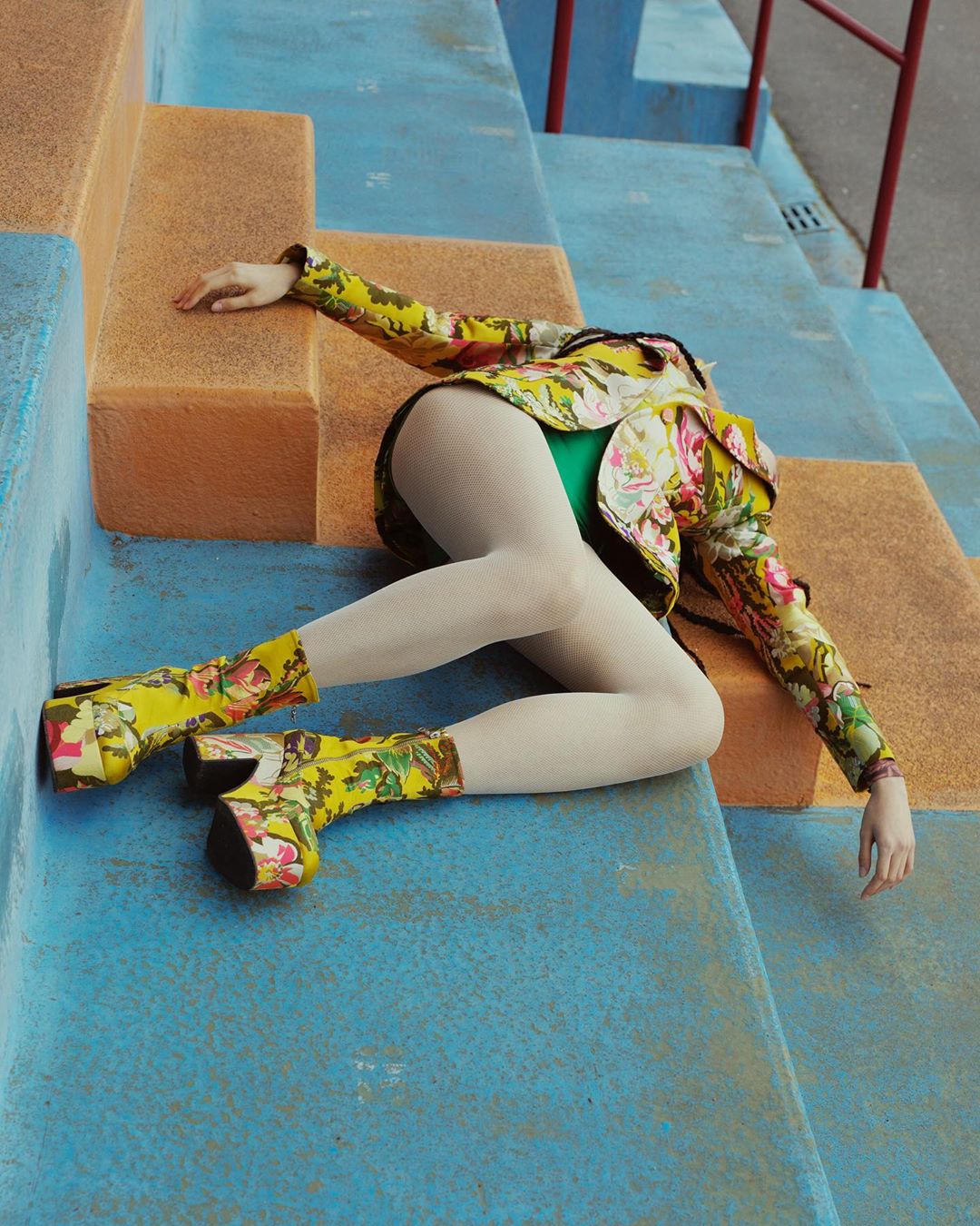 Fashionable woman in floral jacket and boots poses on blue steps, showcasing vibrant colors and patterns in an editorial setting.