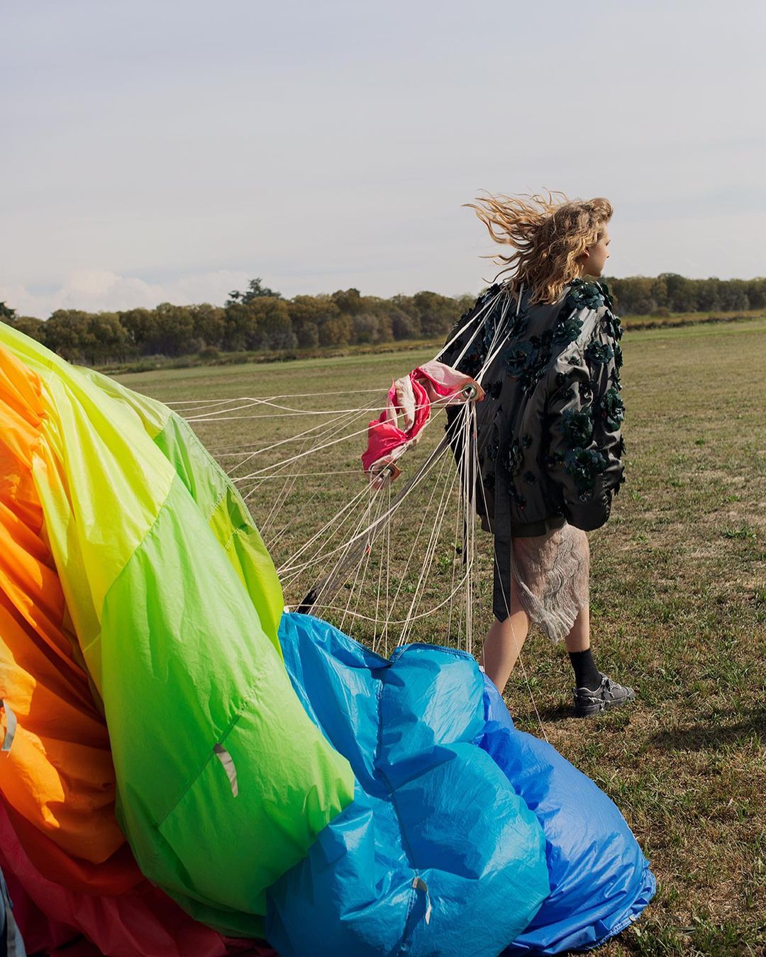 Blonde woman walks in grassy field, dragging deflated rainbow parachute; trees visible in background under lightly clouded sky.