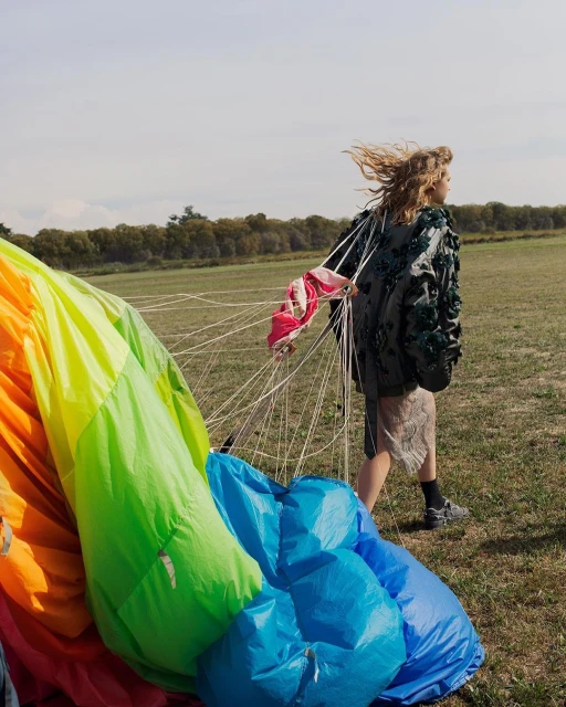Blonde woman walks in grassy field, dragging deflated rainbow parachute; trees visible in background under lightly clouded sky.