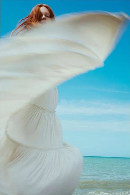 Woman in a billowing white dress stands at the beach, with red hair against a clear blue sky and ocean backdrop.
