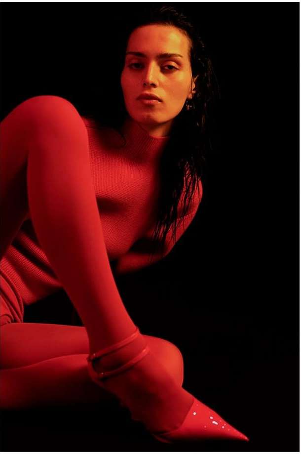 Adult woman poses in a form-fitting red outfit with red high-heels against a dark background in a studio-style setting with dramatic lighting.