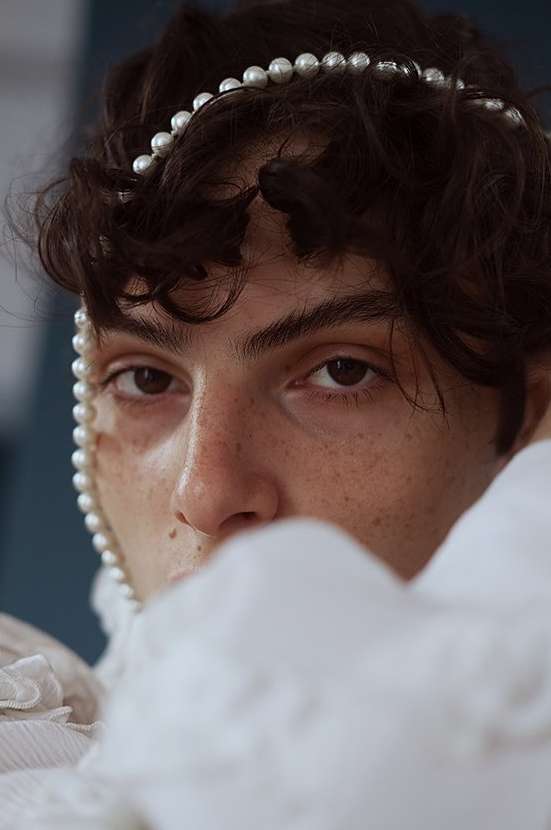 Close-up portrait of a person with dark hair, adorned with a pearl necklace and pearl headband, looking directly at the camera.