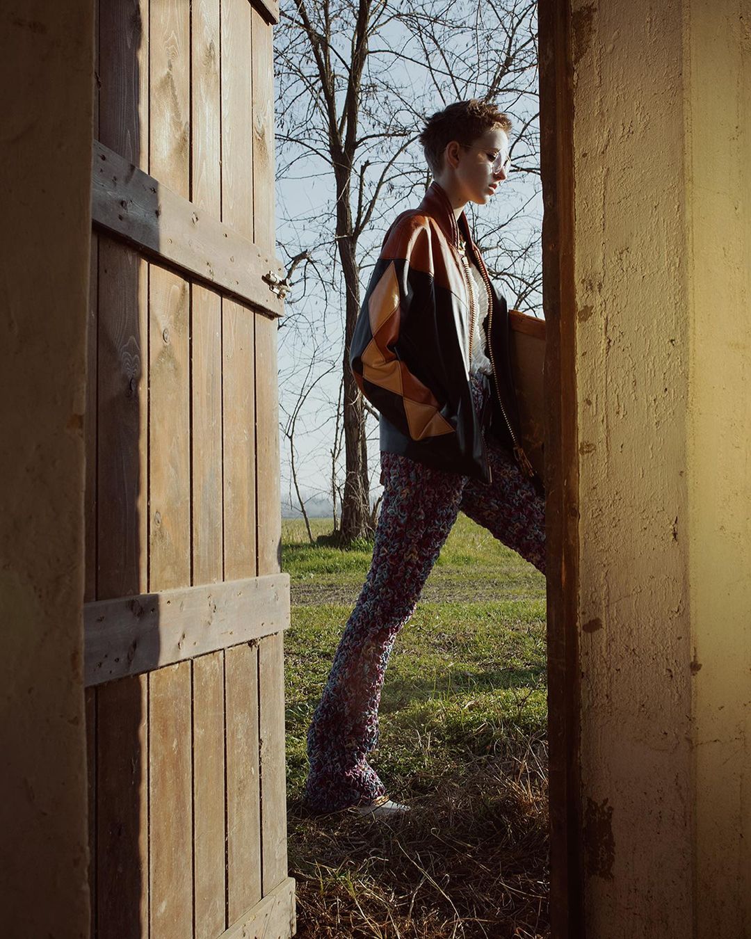 Model with short hair wearing a geometric leather jacket and patterned trousers standing in a wooden doorway in Paris.