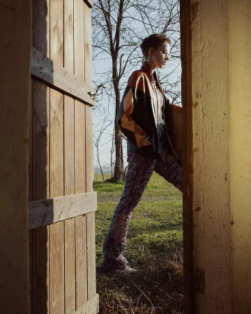 Model with short hair wearing a geometric leather jacket and patterned trousers standing in a wooden doorway in Paris.