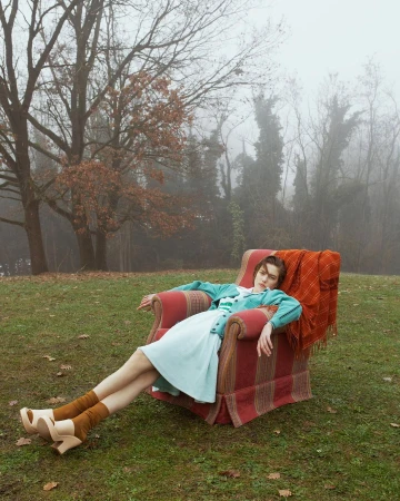 Woman reclining in an armchair on a grassy lawn with bare foggy trees in the background