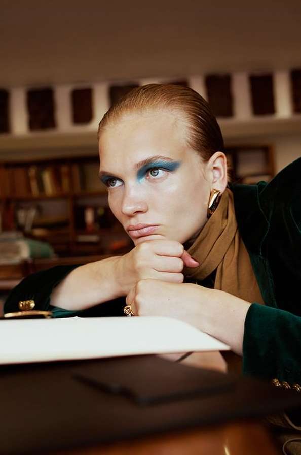 Adult woman with blue eye shadow rests chin on hands while at desk with papers in an indoor environment featuring a bookshelf.