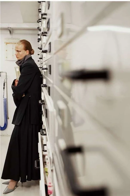 A woman in a black suit stands indoors next to a large set of white drawers, holding a book, in a medium shot.