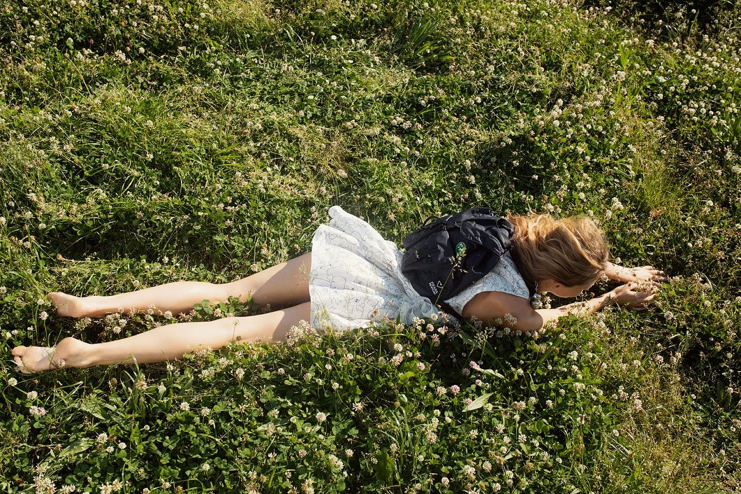 A young woman lying on a lush green field of clover with a backpack, enjoying the sunshine in a peaceful nature setting.