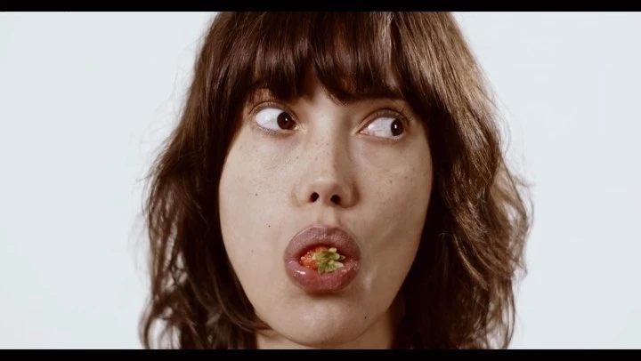 Close-up of a woman with dark hair and fair skin eating a strawberry, set against a plain white background in a studio setting.