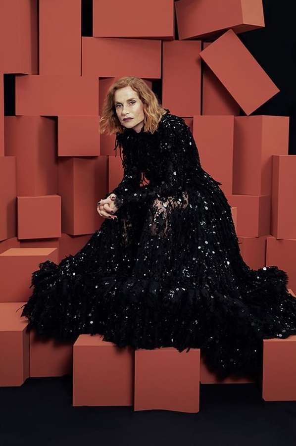 A portrait of an adult female model wearing a long black sequin dress and sitting in front of a geometric block background in a studio setting.