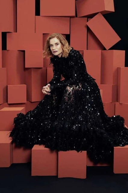 A portrait of an adult female model wearing a long black sequin dress and sitting in front of a geometric block background in a studio setting.