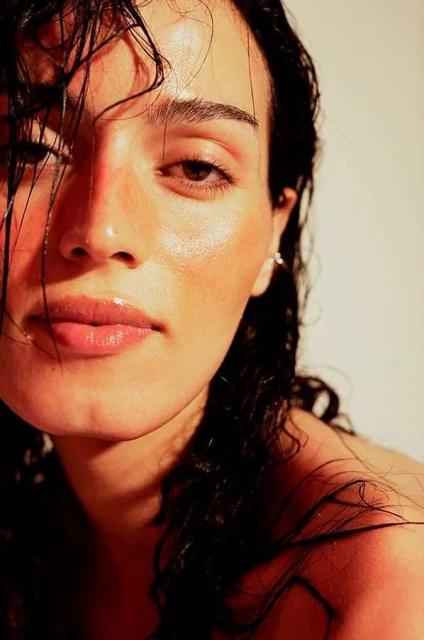Close-up portrait of a young adult female with wet, dark hair, glossy lips, and glowing skin, bathed in warm-toned lighting against a pale background.