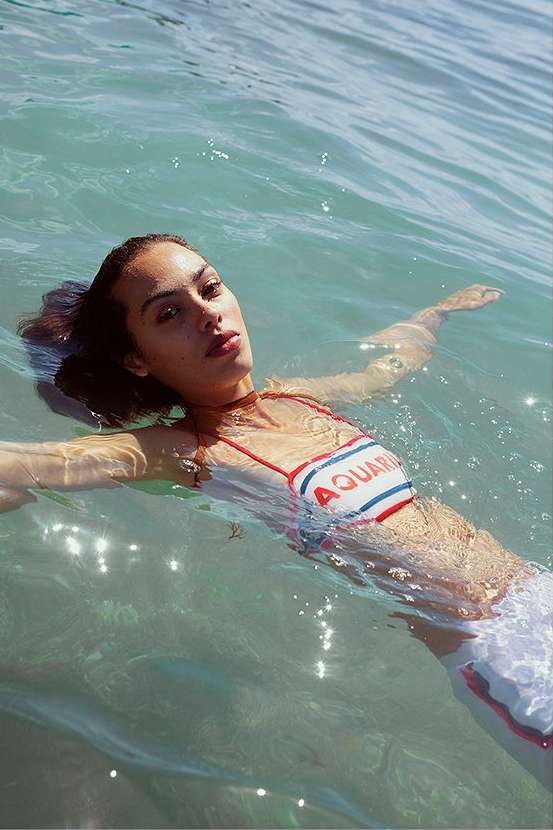 Woman floats on her back in ocean wearing 'Aquarius' top, showcasing swimwear and clear water in an outdoor, summery setting.