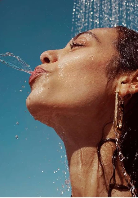 A woman with wet hair drinks water from a shower in front of a blue background. She has earrings and water is splashing on her face.