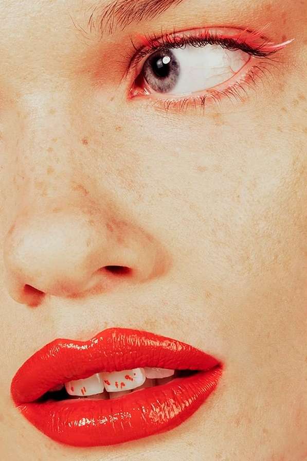 Close-up portrait of a woman's face featuring vibrant red lipstick, freckled skin, and detailed eye makeup, showcasing beauty and detail.