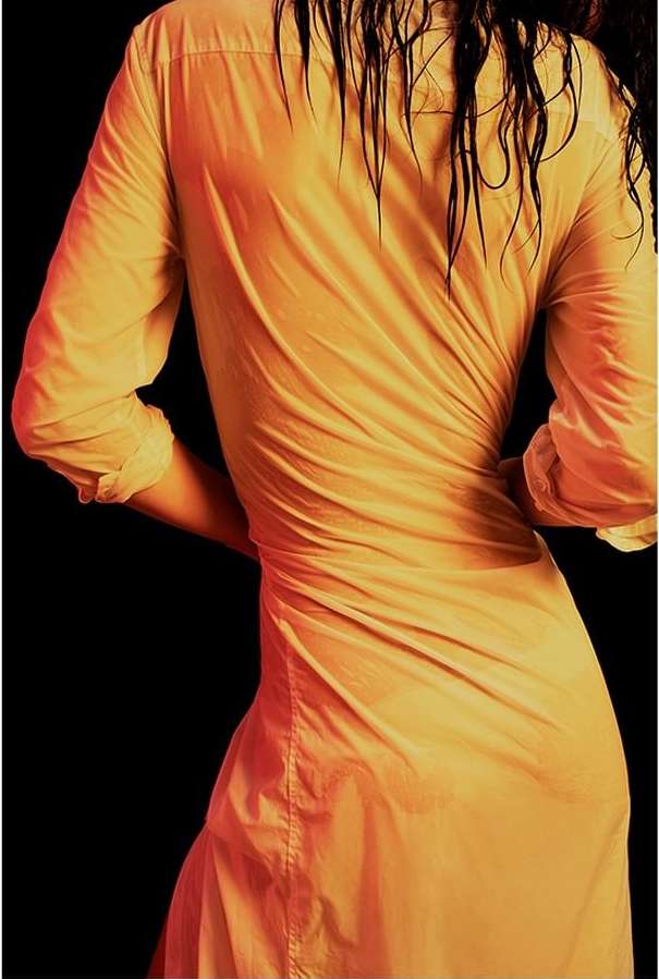 A woman with wet hair stands with her back to the camera, wearing a yellow shirt dress against a black background in a studio setting.
