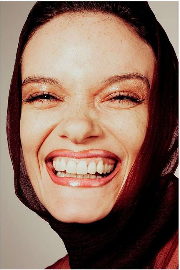 Close-up portrait of a smiling woman with brown hair, freckles, and a translucent brown headscarf against a light grey background.