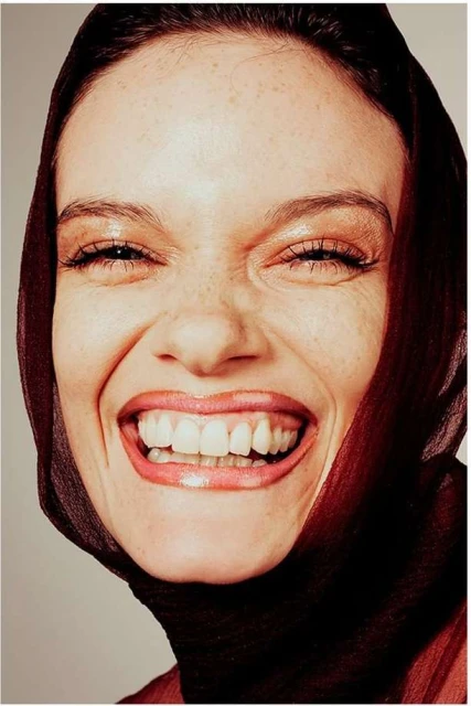 Close-up portrait of a smiling woman with brown hair, freckles, and a translucent brown headscarf against a light grey background.