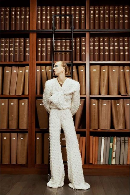Stylish woman in white suit posing near bookshelf with ladder in library. Fashion editorial shot of model looking off camera.