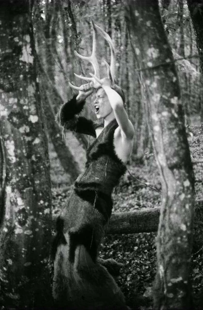 Person in furry costume and large antlers stands in a forest, mouth open, in black and white. Trees surround the subject.