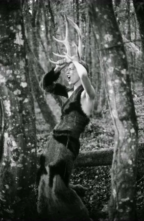 Person in furry costume and large antlers stands in a forest, mouth open, in black and white. Trees surround the subject.