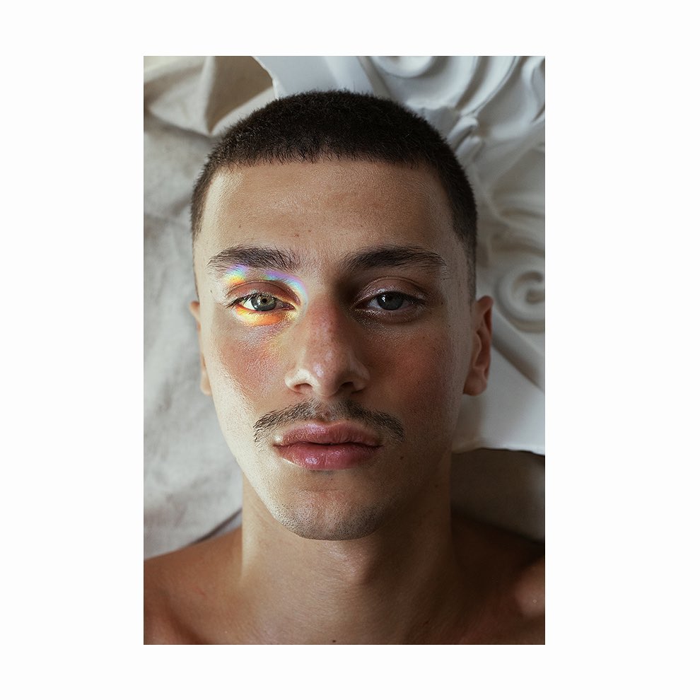 Close-up portrait of a fair-skinned adult male with a mustache, short dark hair, and a rainbow light effect on his face, indoors.