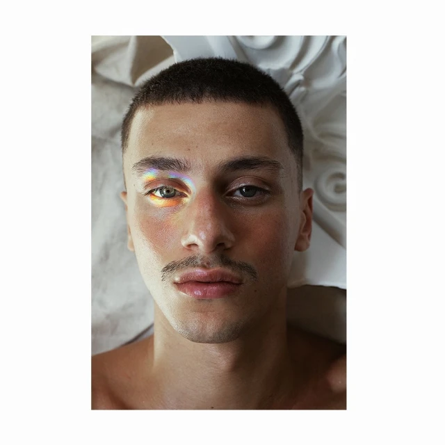 Close-up portrait of a fair-skinned adult male with a mustache, short dark hair, and a rainbow light effect on his face, indoors.