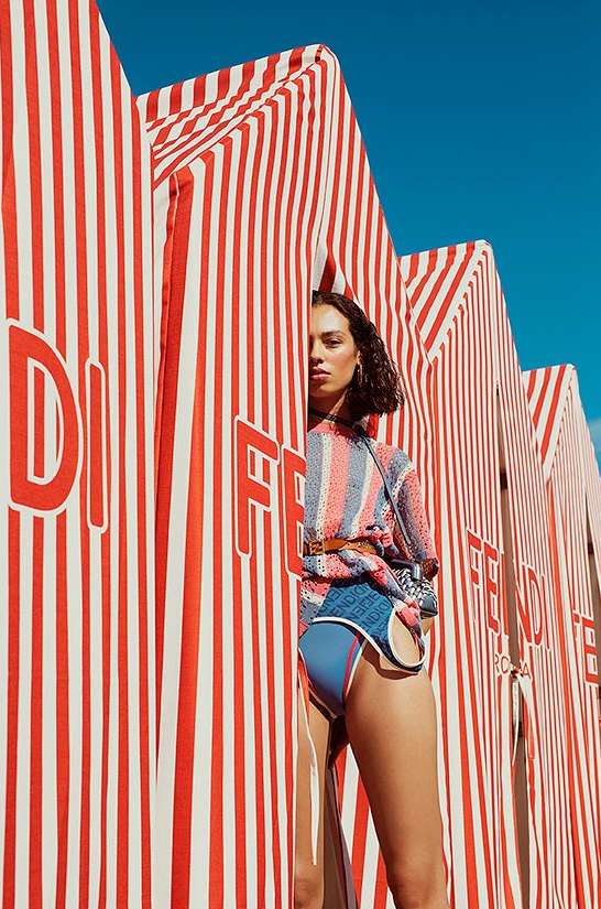 A woman posing in a blue and orange swimsuit stands between striped structures on a bright day, showcasing fashion and summer vibes.