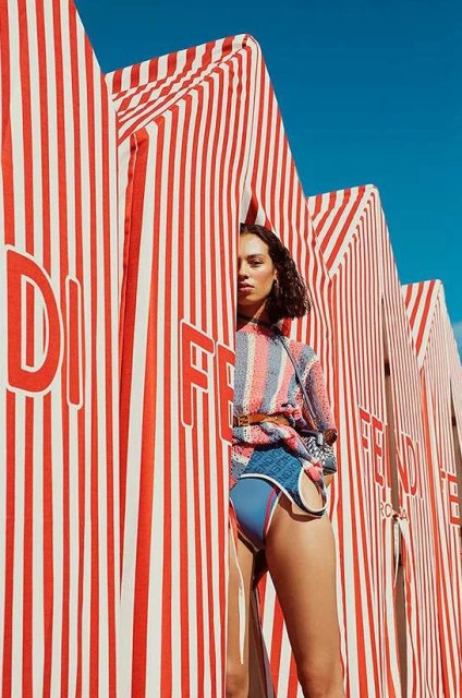 A woman posing in a blue and orange swimsuit stands between striped structures on a bright day, showcasing fashion and summer vibes.