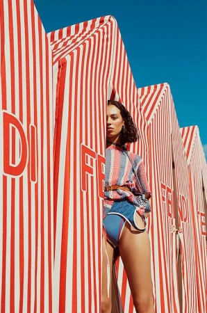 A woman posing in a blue and orange swimsuit stands between striped structures on a bright day, showcasing fashion and summer vibes.