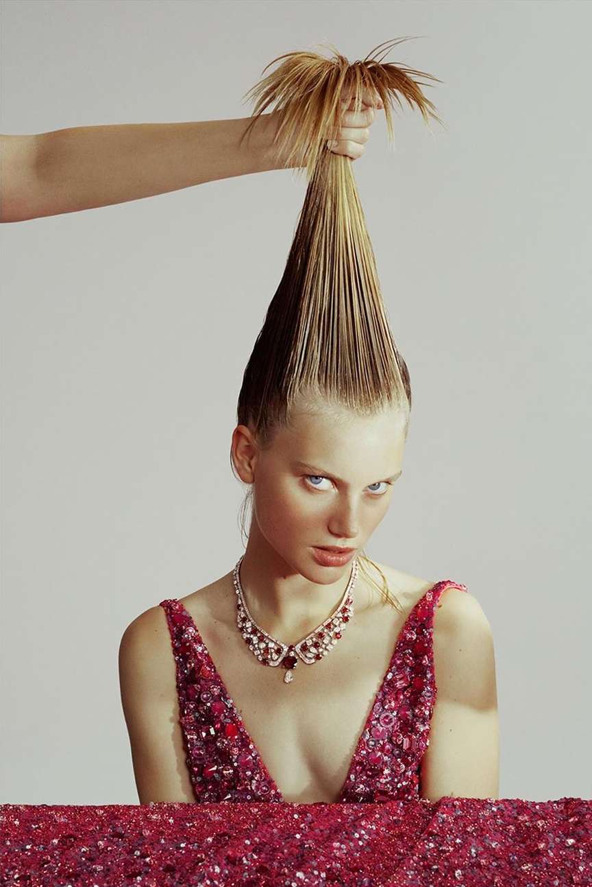 Woman in a pink dress with a ruby necklace has her blonde hair held up in a point on a neutral background, high-fashion editorial.