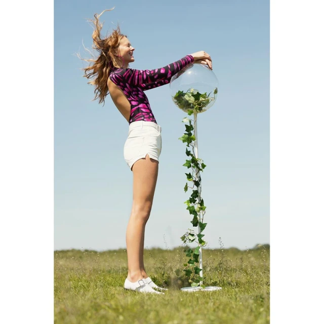 Adult female stands outside in open grassy field next to a large clear plastic ball filled with greenery and ivy, under clear skies.