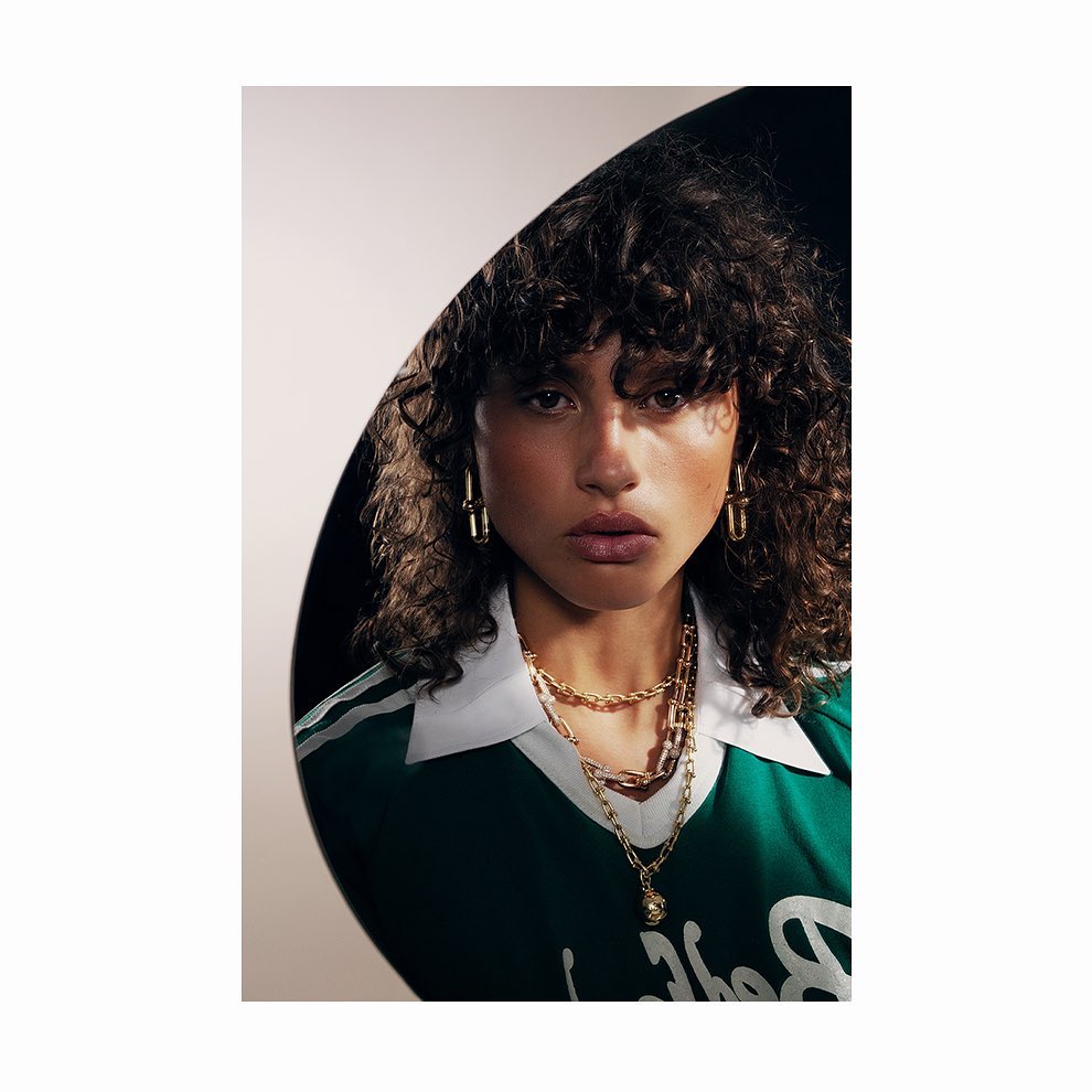 Close-up studio shot of a young adult woman with curly hair wearing gold earrings and gold necklaces, posing against a dark backdrop.