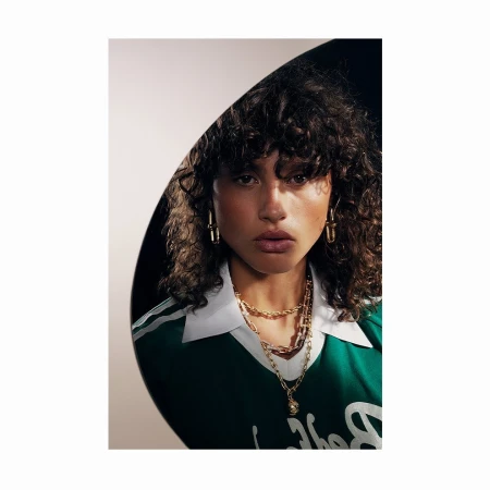 Close-up studio shot of a young adult woman with curly hair wearing gold earrings and gold necklaces, posing against a dark backdrop.