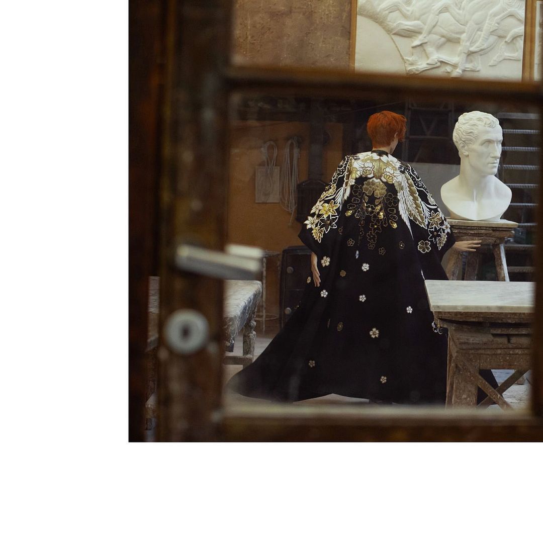 Back view of a person wearing a black floral robe standing near a classical bust in a room with classical-style architecture, viewed through a window.