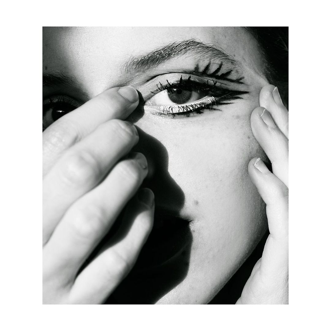 Close-up black-and-white portrait of a person with stylized graphic eye makeup, skin details, and partial face and hands in view.
