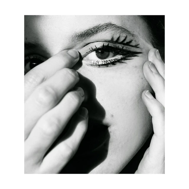 Close-up black-and-white portrait of a person with stylized graphic eye makeup, skin details, and partial face and hands in view.