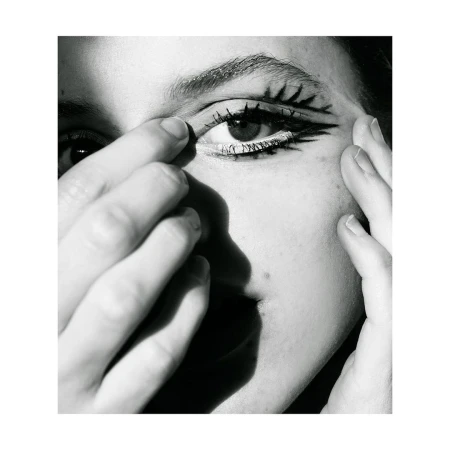 Close-up black-and-white portrait of a person with stylized graphic eye makeup, skin details, and partial face and hands in view.
