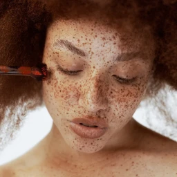 Close-up portrait of a woman with dense freckles and curly hair being applied with red makeup by a brush in Paris.