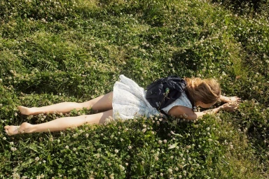 Woman wearing a patterned white dress and dark backpack lying prone in a lush green meadow filled with white clover flowers.