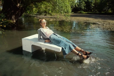 A laughing woman lying on a white desk submerged in a shallow river, wearing a grey blazer, patterned top, and denim skirt.